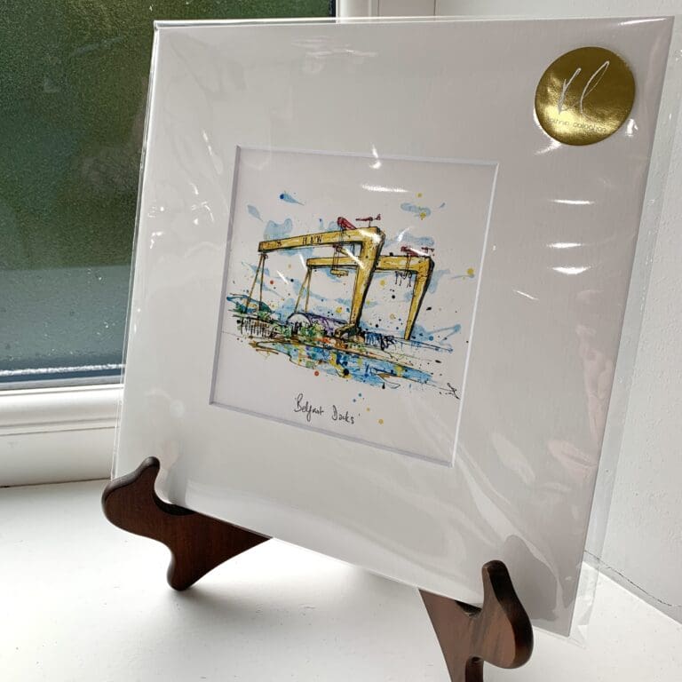 A miniature print of Belfast Docks by Kathryn Callaghan, which features a landscape image of Belfast's Harland & Wolff yellow cranes. The print is presented mounted at 23cm, in a clear cello bag with a gold KC sticker in the top right corner, and is displayed here on a small wooden easel in a windowsill.