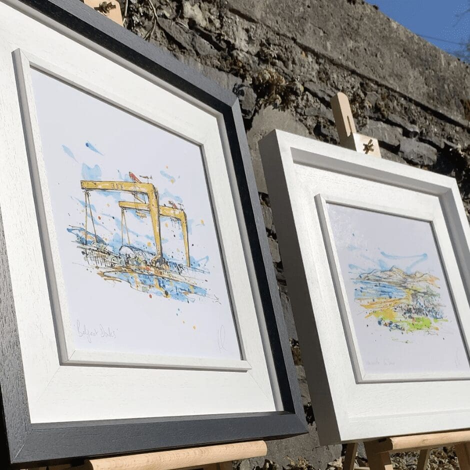 Two prints by Kathryn Callaghan photographed in frames on small easels outdoors. The prints are Belfast Docks, which features a view of the yellow Harland & Wolff cranes in Belfast, in a two part white and black frame, and Newcastle Co. Down, which features a view of the Mourne Mountains and Newcastle in a matching white frame.