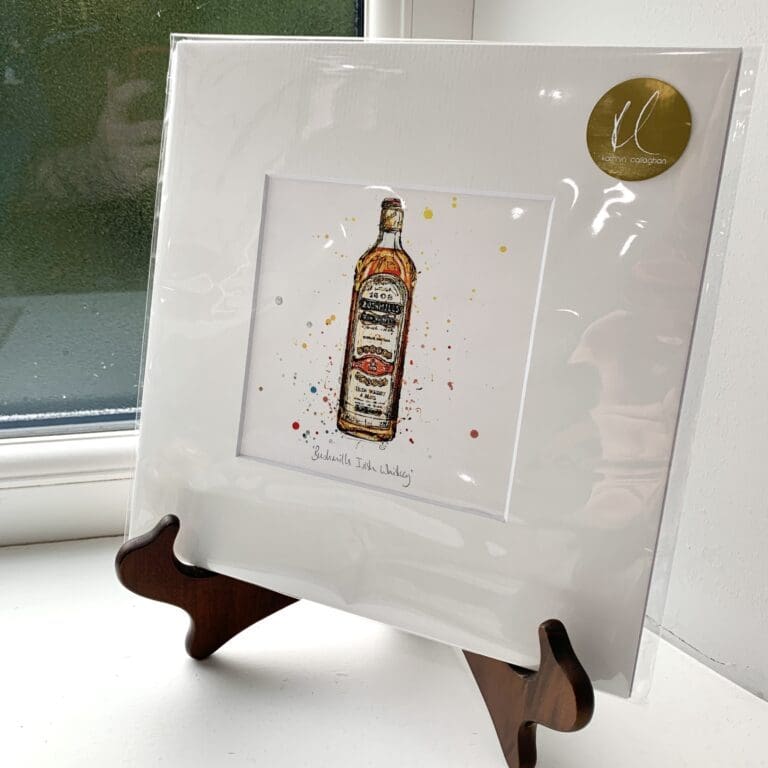 A miniature print of Bushmills Irish Whiskey by Kathryn Callaghan, which features the eponymous golden bottle with its iconic label. The print is presented mounted at 23cm, in a clear cello bag with a gold KC sticker in the top right corner, and is displayed here on a small wooden easel in a windowsill.