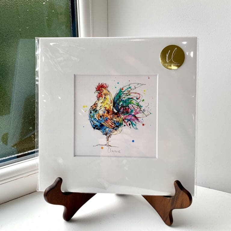 A miniature print of Clarence by Kathryn Callaghan, which features a colourful cockerel facing left. The print is presented mounted at 23cm, in a clear cello bag with a gold KC sticker in the top right corner, and is displayed here on a small wooden easel in a windowsill.