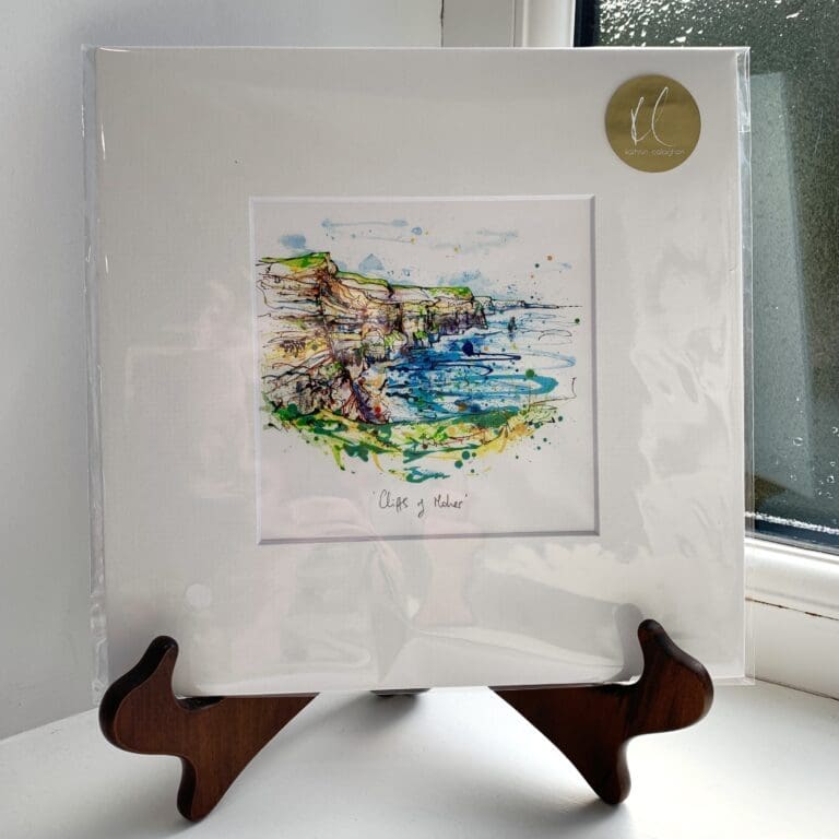 A miniature print of Cliffs of Moher by Kathryn Callaghan, which features the eponymous Irish cliffs rising high above the bright blue Atlantic. The print is presented mounted at 23cm, in a clear cello bag with a gold KC sticker in the top right corner, and is displayed here on a small wooden easel in a windowsill.