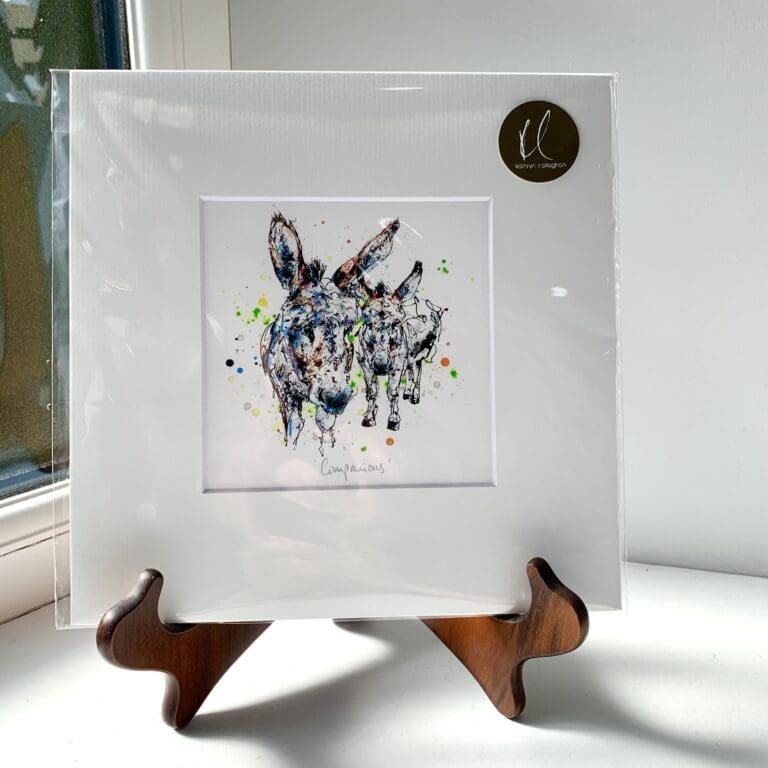 A miniature print of Companions by Kathryn Callaghan, which features two donkeys looking curiously at the viewer. The print is presented mounted at 23cm, in a clear cello bag with a gold KC sticker in the top right corner, and is displayed here on a small wooden easel in a windowsill.