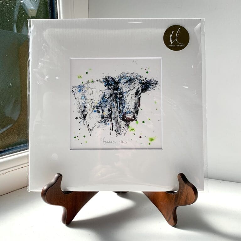 A miniature print of Pastures New by Kathryn Callaghan, which features a black and white dairy cow looking at the viewer. The print is presented mounted at 23cm, in a clear cello bag with a gold KC sticker in the top right corner, and is displayed here on a small wooden easel in a windowsill.