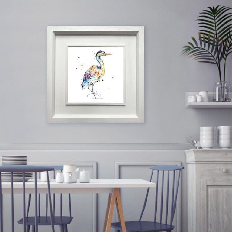 A print of a standing heron named Serenity by Kathryn Callaghan, shown framed in a deluxe white frame in situ in a white kitchen with powder blue chairs.