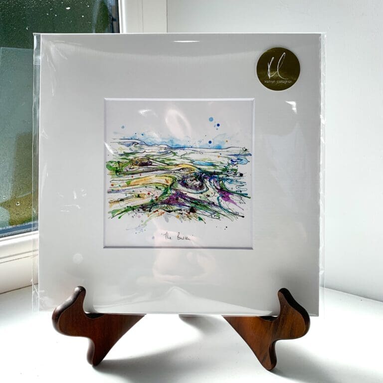 A miniature print of The Burren by Kathryn Callaghan, which features the eponymous Irish landscape. The print is presented mounted at 23cm, in a clear cello bag with a gold KC sticker in the top right corner, and is displayed here on a small wooden easel in a windowsill.