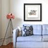 A 30x30cm print by Kathryn Callaghan shown in a deluxe black frame in situ above a lilac sofa and pink floral lamp. The print is Well, hello, which shows three dairy cows looking over a red fence gate.