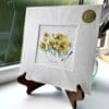 A miniature print of Happy Days by Kathryn Callaghan, which features a vivid spray of yellow daffodils. The print is presented mounted at 23cm, in a clear cello bag with a gold KC sticker in the top right corner, and is displayed here on a small wooden easel in a windowsill.