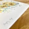 Photograph of Happy Days by Kathryn Callaghan, a print of a vivid yellow spray of daffodils, which shows the print's title handwritten in the bottom left corner and the artist's signature in the bottom right, with space between to add a custom personalisation.