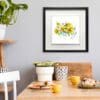 'Happy Days', a vivid daffodil print by Kathryn Callaghan, shown displayed in a Deluxe Black Frame in situ above a sunny breakfast table laid with cups of coffee and bread.