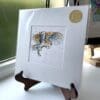 A miniature print of Hover by Kathryn Callaghan, which features a barn owl in flight. The print is presented mounted at 23cm, in a clear cello bag with a gold KC sticker in the top right corner, and is displayed here on a small wooden easel in a windowsill.