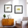 Two prints by Kathryn Callaghan displayed in situ in an airy lounge with white walls, a teak sideboard, and a comfortable white armchair. The prints are Hover, which features a barn owl in flight, and Happy Days, which features a spray of butter-yellow daffodils. Both prints are framed in Deluxe two-part Black Frames.