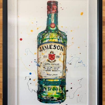 An original painting of a bottle of Jameson Irish Whiskey by Kathryn Callaghan, shown flat on a wooden background. The painting is framed in a two part white inner and black outer wood frame.
