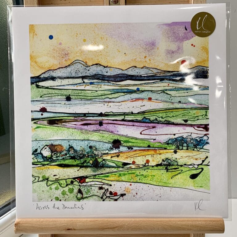 A print of Across the Drumlins by Kathryn Callaghan, which features a view across the rolling landscape to the Mourne Mountains. The 30cm print is presented flat in a clear cello bag with a golden KC sticker in the top right.