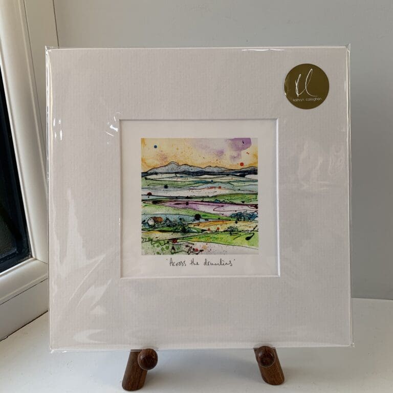 A miniature print of Across the Drumlins by Kathryn Callaghan, which features a landscape of rolling hills. The print is presented mounted at 23cm, in a clear cello bag with a gold KC sticker in the top right corner, and is displayed here on a small wooden easel in a windowsill.