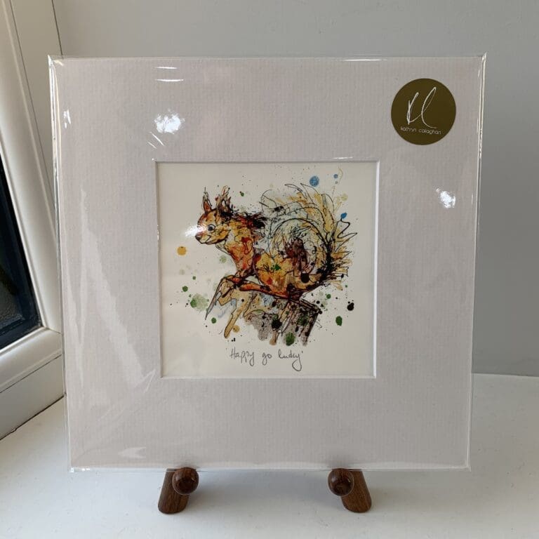 A miniature print of Happy Go Lucky by Kathryn Callaghan, which features a red squirrel perched on a stump. The print is presented mounted at 23cm, in a clear cello bag with a gold KC sticker in the top right corner, and is displayed here on a small wooden easel in a windowsill.