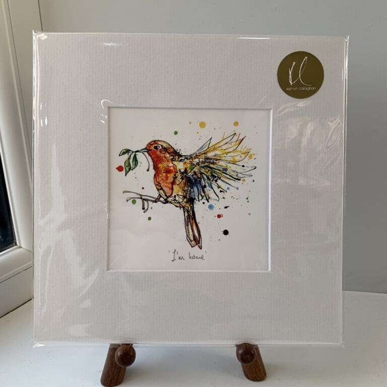 A miniature print of I'm Home by Kathryn Callaghan, which features a robin in flight holding a sprig of mistletoe. The print is presented mounted at 23cm, in a clear cello bag with a gold KC sticker in the top right corner, and is displayed here on a small wooden easel in a windowsill.