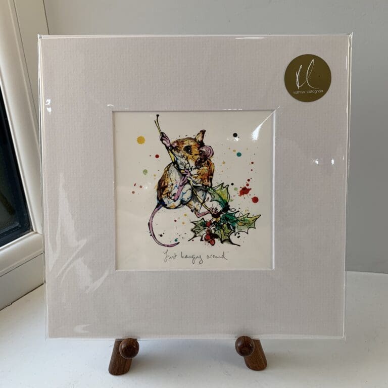 A miniature print of Just Hanging Around by Kathryn Callaghan, which features a harvest mouse clinging to a sprig of holly. The print is presented mounted at 23cm, in a clear cello bag with a gold KC sticker in the top right corner, and is displayed here on a small wooden easel in a windowsill.