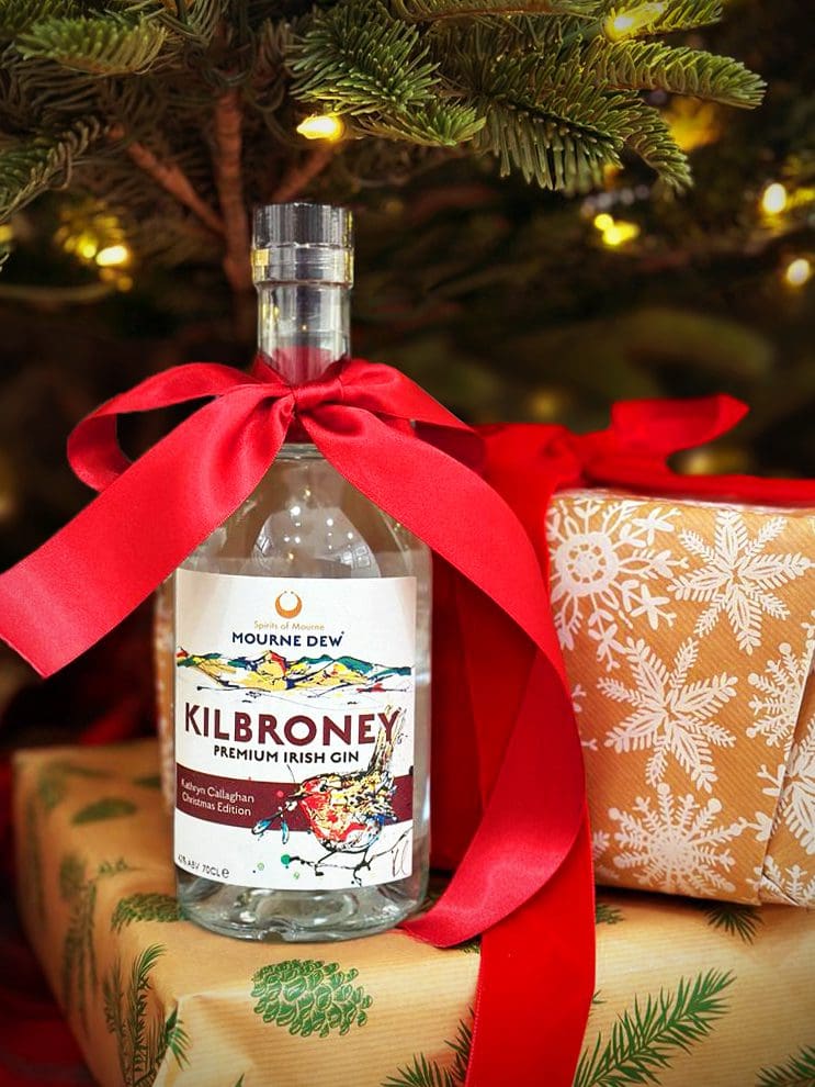 A bottle of Mourne Dew Kilbroney gin sporting a red velvet ribbon tied in a bow, placed atop two wrapped Christmas presents below a Christmas tree. The bottle's label reads "Mourne Dew Kilbroney Premium Irish Gin", and features a painted robin by Kathryn Callaghan alongside text reading "Kathryn Callaghan Christmas Edition."