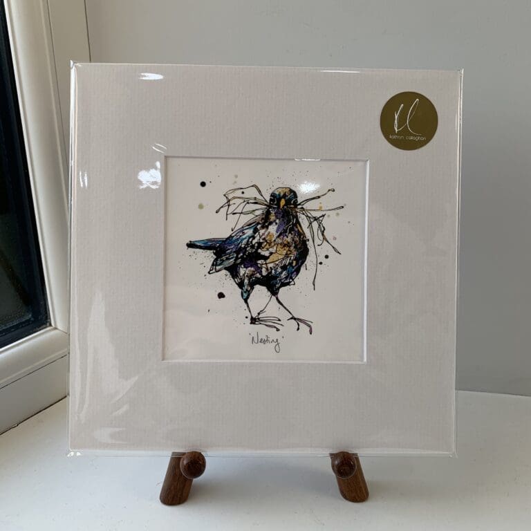 A miniature print of Nesting by Kathryn Callaghan, which features a blackbird with a beak full of straw. The print is presented mounted at 23cm, in a clear cello bag with a gold KC sticker in the top right corner, and is displayed here on a small wooden easel in a windowsill.