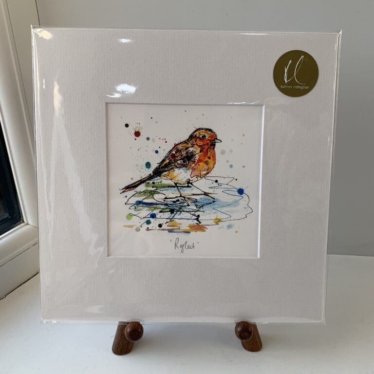 A miniature print of Reflect by Kathryn Callaghan, which features a robin standing in a small pool of water. The print is presented mounted at 23cm, in a clear cello bag with a gold KC sticker in the top right corner, and is displayed here on a small wooden easel in a windowsill.