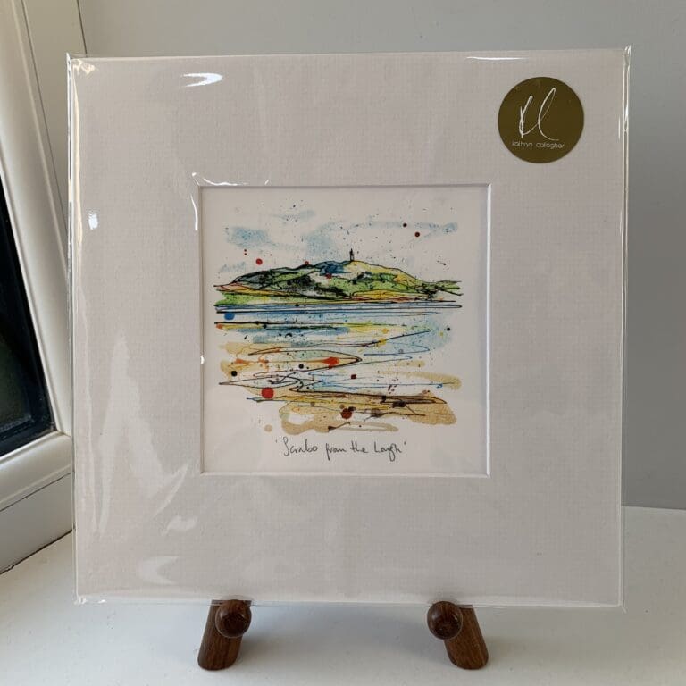 A miniature print of Scrabo From The Lough by Kathryn Callaghan, which features a view of Scrabo tower across Strangford Lough. The print is presented mounted at 23cm, in a clear cello bag with a gold KC sticker in the top right corner, and is displayed here on a small wooden easel in a windowsill.