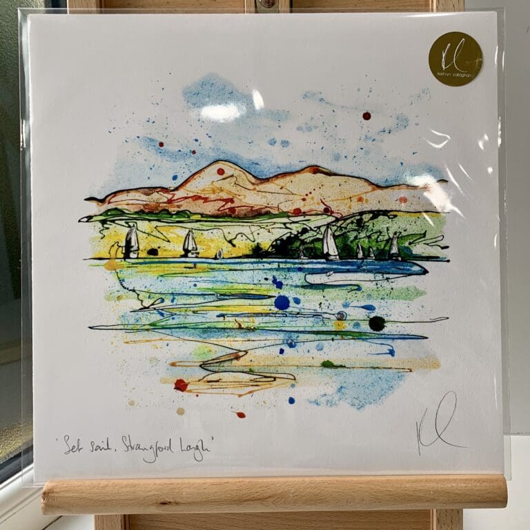 A print of Set Sail, Strangford Lough by Kathryn Callaghan, which features a view of the Mourne Mountains from Strangford Lough with sailboats on the water. The 30cm print is presented flat in a clear cello bag with a golden KC sticker in the top right.