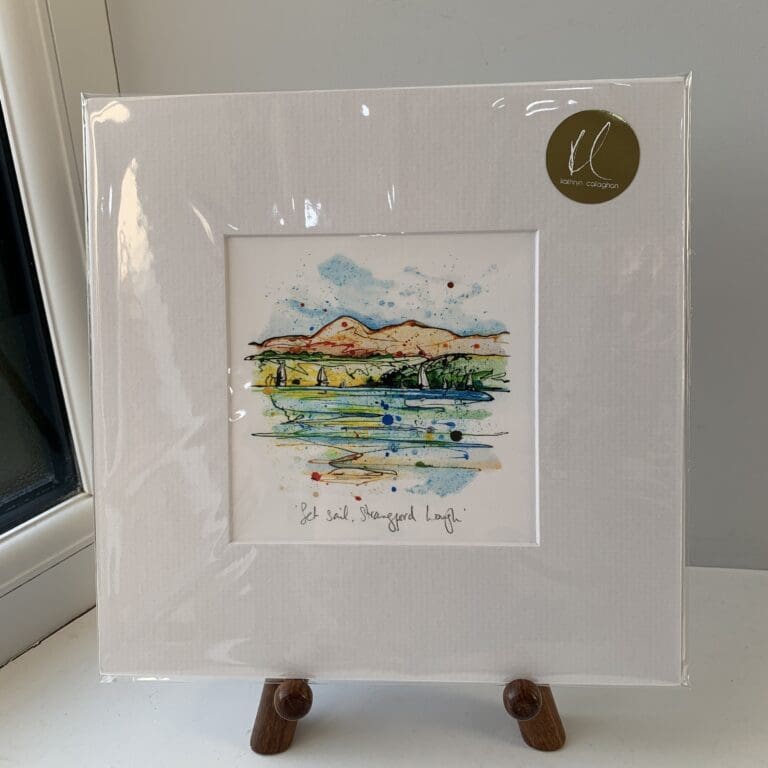 A miniature print of Set Sail, Strangford Lough by Kathryn Callaghan, which features a view across Strangford Lough to the Mournes with sailboats on the water. The print is presented mounted at 23cm, in a clear cello bag with a gold KC sticker in the top right corner, and is displayed here on a small wooden easel in a windowsill.