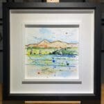 An original painting named Set Sail, Strangford Lough by Kathryn Callaghan, featuring a view from the surface of Strangford Lough to the Mournes in Northern Ireland, with sailboats on the water. The painting is framed in a two part white and black frame.