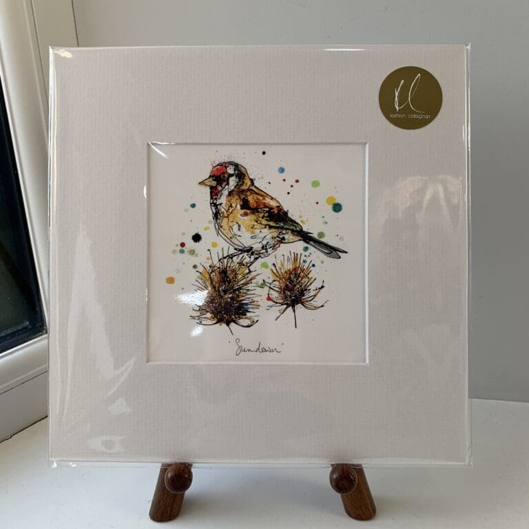 A miniature print of Sundown by Kathryn Callaghan, which features a goldfinch perched on a thistle. The print is presented mounted at 23cm, in a clear cello bag with a gold KC sticker in the top right corner, and is displayed here on a small wooden easel in a windowsill.