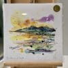 A print of The Mournes at Sunset by Kathryn Callaghan, which features a view of the Mourne Mountains with a purple and golden sunset in the background. The 30cm print is presented flat in a clear cello bag with a golden KC sticker in the top right.