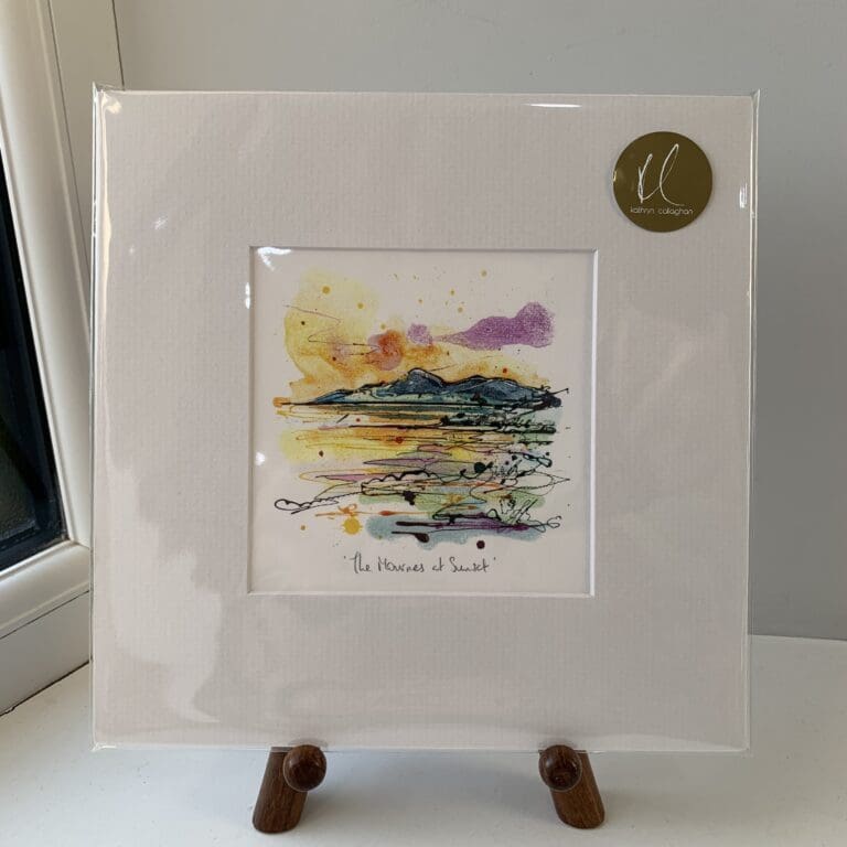 A miniature print of The Mournes at Sunset by Kathryn Callaghan, which features a view of The Mourne Mountains with a purple and golden sunset in the background. The print is presented mounted at 23cm, in a clear cello bag with a gold KC sticker in the top right corner, and is displayed here on a small wooden easel in a windowsill.