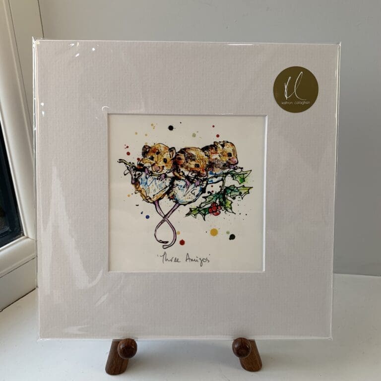 A miniature print of Three Amigos by Kathryn Callaghan, which features three harvest mice clinging to a holly twig. The print is presented mounted at 23cm, in a clear cello bag with a gold KC sticker in the top right corner, and is displayed here on a small wooden easel in a windowsill.