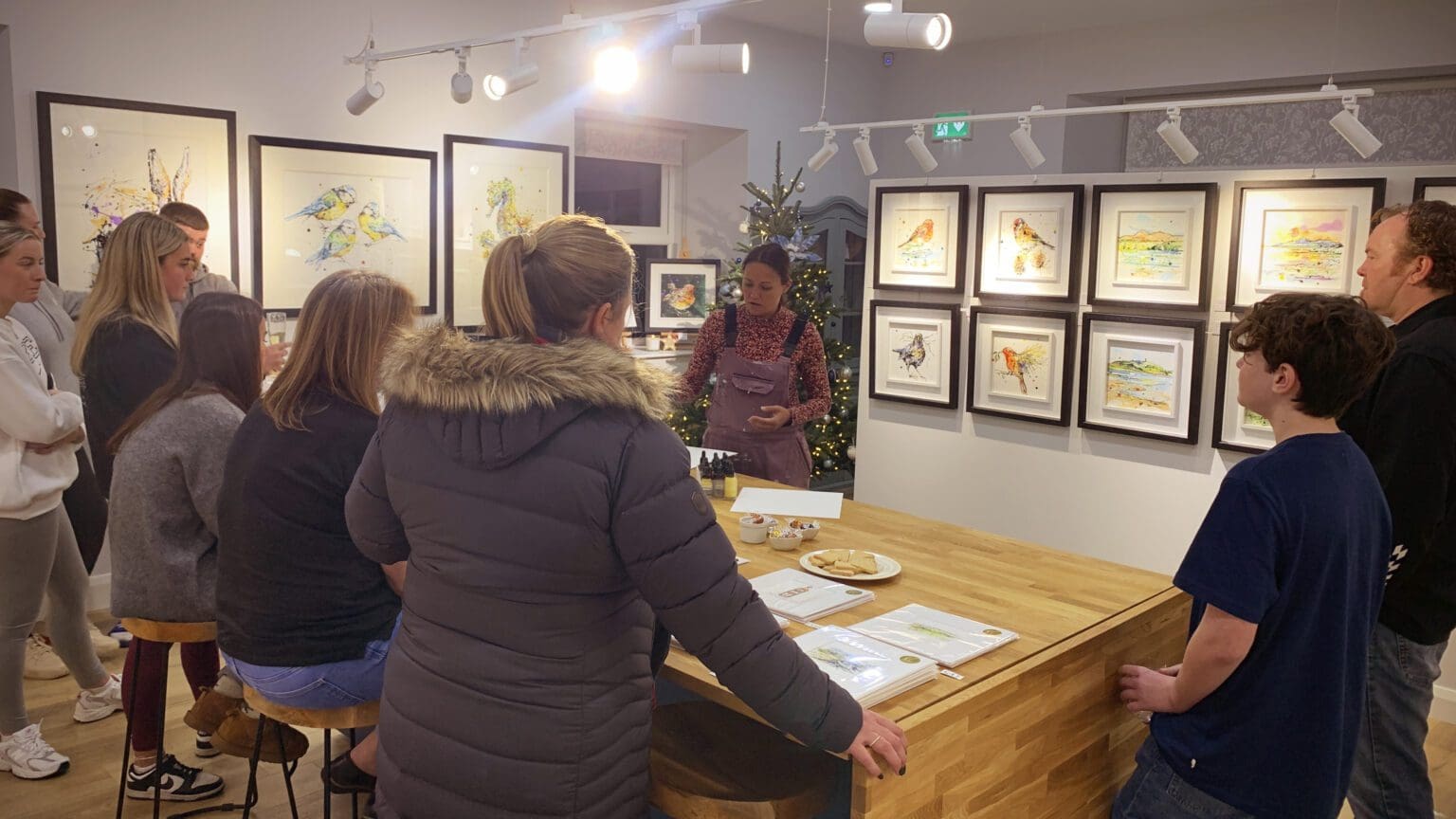 Guests in the Kathryn Callaghan Fine Art Studio, stood around a wooden island, watching Kathryn Callaghan demonstrate her painting technique. New original paintings line the walls.