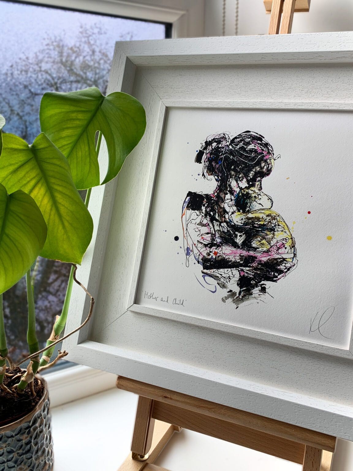 A print of Mother And Child by Kathryn Callaghan, displayed in a Deluxe White Frame on an easel alongside a monstera plant.