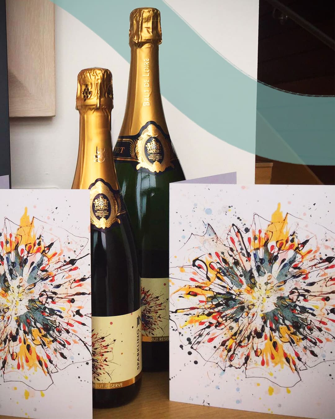 Two bottles of JN Wine Sparkling Brut Reserve sat on a table with two folded cards bearing the same artwork, which features an abstract flower painted similarly to a firework by Kathryn Callaghan.