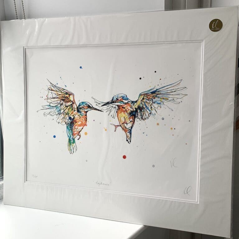 A mounted print of a pair of kingfishers in flight by Kathryn Callaghan, named Togetherness. The print is shown upright in a sunny windowsill.