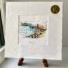 A miniature print of Bangor Pier by Kathryn Callaghan, which features the County Down city's pier, harbour and lighthouse. The print is presented mounted at 23cm, in a clear cello bag with a gold KC sticker in the top right corner, and is displayed here on a small wooden easel in a windowsill.