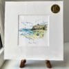 A miniature print of Benone Strand by Kathryn Callaghan, which features Benone beach and cliffs with Mussenden Temple sat atop. The print is presented mounted at 23cm, in a clear cello bag with a gold KC sticker in the top right corner, and is displayed here on a small wooden easel in a windowsill.