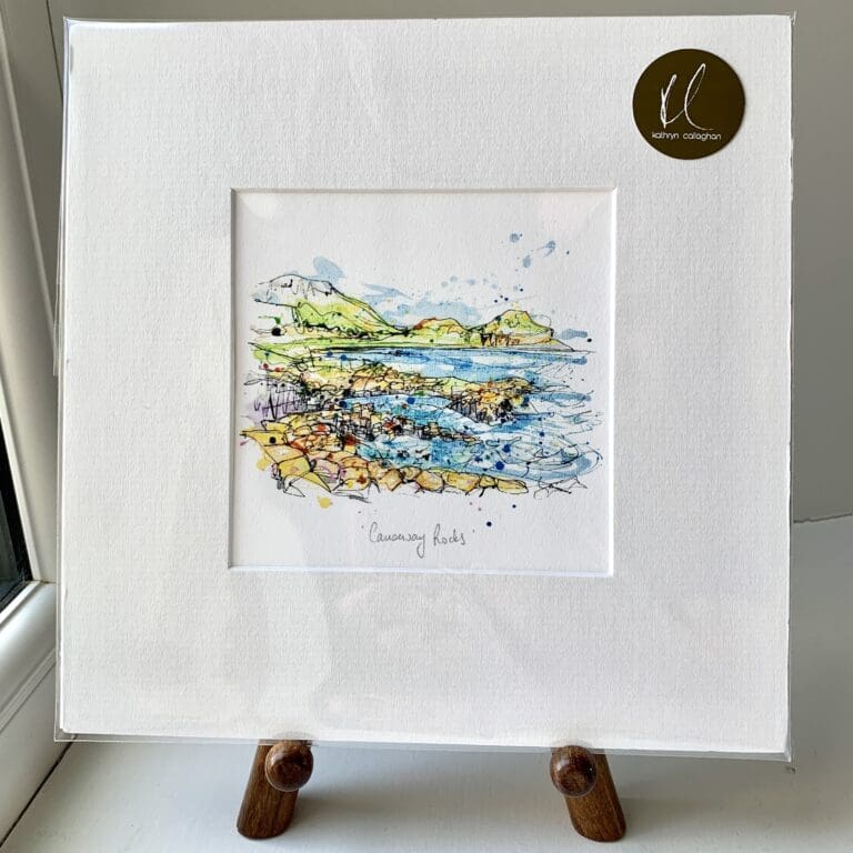 A miniature print of Causeway Rocks by Kathryn Callaghan, which features the Giants Causeway. The print is presented mounted at 23cm, in a clear cello bag with a gold KC sticker in the top right corner, and is displayed here on a small wooden easel in a windowsill.