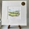 A miniature print of Glens of Antrim by Kathryn Callaghan, which features the rolling County Antrim hills. The print is presented mounted at 23cm, in a clear cello bag with a gold KC sticker in the top right corner, and is displayed here on a small wooden easel in a windowsill.